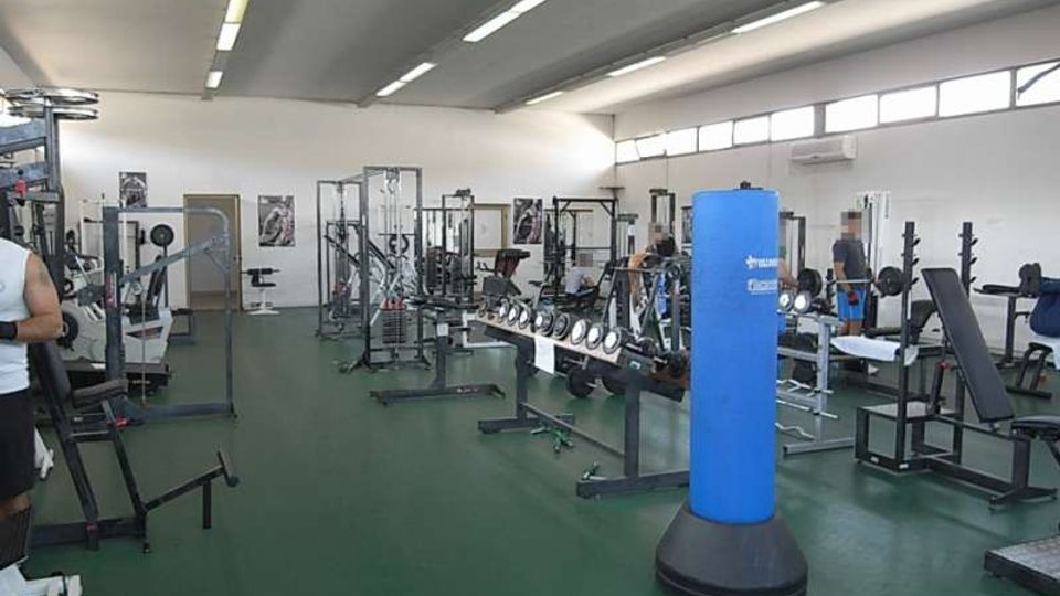 Interno di una palestra con ampio open space, pavimento verde e soffitto alto con luci lineari. Sono presenti panche e rack per pesi, una lunga rastrelliera di manubri e un sacco da boxe blu al centro, con grandi finestre alte lungo una parete.