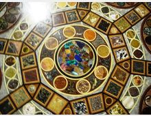 Dettaglio ravvicinato di un soffitto a cupola decorato con mosaici geometrici, con un medaglione circolare centrale in vetro colorato e anelli concentrici di riquadri e cerchi con motivi ornamentali. La superficie è suddivisa in pannelli multicolori incorniciati, con una forte luce proveniente da un lato.