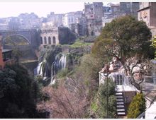 Veduta panoramica di un borgo storico su una gola, con edifici in pietra, un ponte ad arco e un antico viadotto sullo sfondo. In basso si vede una cascata che scende tra vegetazione fitta, con terrazze e scale lungo il versante sulla destra.