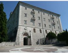 Grande edificio in pietra chiara a più piani, con piccole finestre regolari e balconcini sporgenti, visto da un piazzale lastricato con motivi geometrici. Al piano terra si nota un portale ad arco con la scritta “PAX”, un cancello laterale e alberi ai margini.