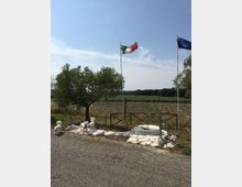 Area rurale con campo coltivato e filari sullo sfondo, delimitata da una staccionata in legno e pietre bianche vicino a una strada asfaltata. Due alti pennoni con bandiere italiana e dell’Unione Europea si trovano accanto a un ulivo e a una piccola aiuola circolare.
