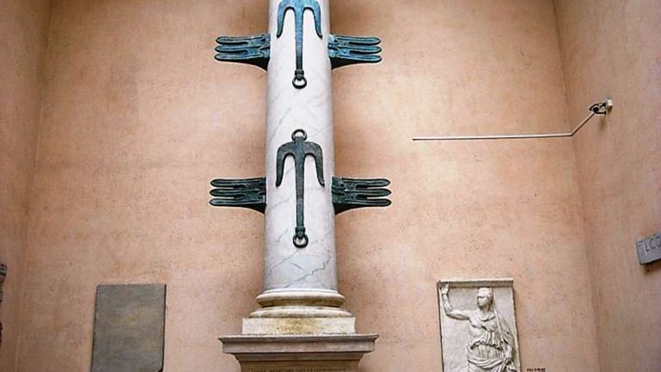 Sala museale con pareti color ocra e lucernario, dominata da una colonna marmorea alta con elementi metallici decorativi sporgenti. In primo piano è esposto un lungo reperto lapideo su supporti, con bassorilievi e targhe informative alle pareti.