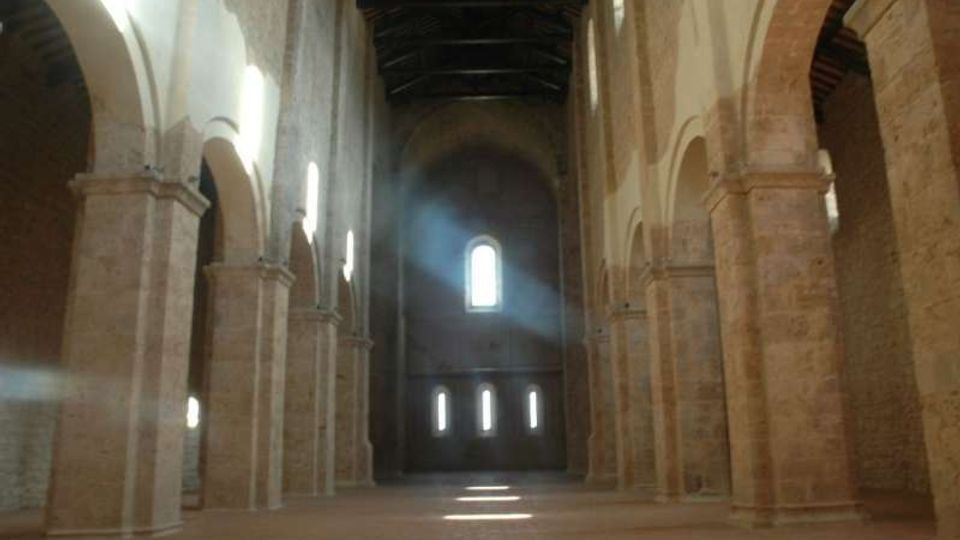 Interno di una chiesa in stile romanico con navata centrale ampia, file di pilastri in pietra e arcate laterali. Piccole finestre ad arco sul fondo lasciano entrare fasci di luce che illuminano il pavimento.