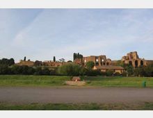 Panorama di rovine archeologiche in mattoni su una leggera altura, con archi e murature antiche tra alberi alti e vegetazione. In primo piano si vede un ampio prato con un sentiero asfaltato e cielo sereno sullo sfondo.
