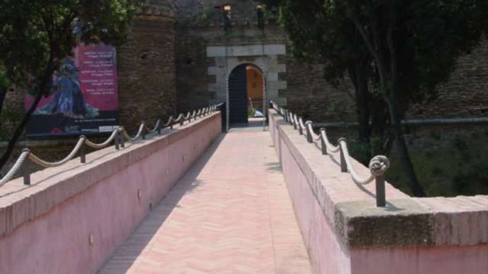 Passerella pedonale in mattoni con parapetti intonacati e catene, che conduce a un portale ad arco in pietra su un muro di fortificazione in mattoni. Ai lati si vedono alberi e un grande cartellone, con vegetazione che circonda l’ingresso.
