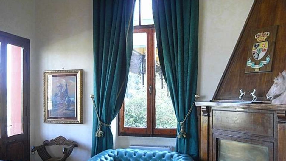 Salotto con divani Chesterfield in pelle azzurra, tavolino in legno su tappeto decorato e grande finestra con tende pesanti verde scuro. A destra c’è un camino rivestito in legno con cappa triangolare e stemma, con soffitto in legno e pareti chiare.