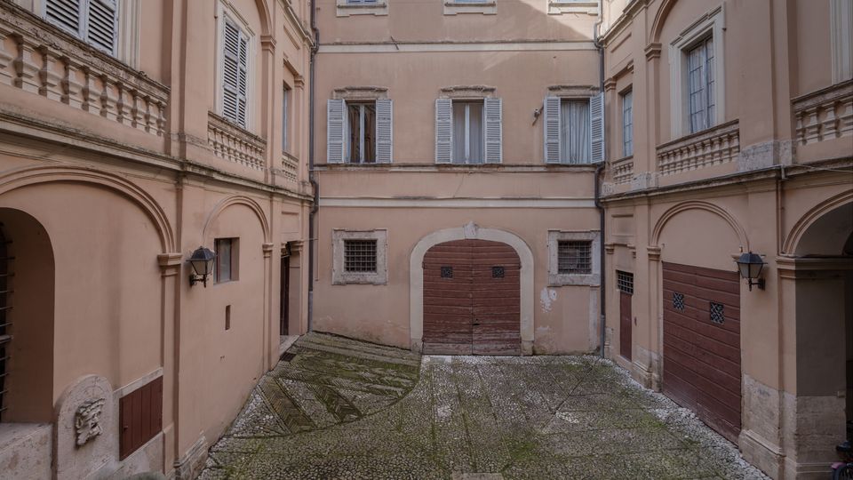 Cortile interno di un edificio storico con facciate color beige, archi e finestre con persiane. Al centro si vede un grande portone in legno e una pavimentazione in ciottoli con motivi geometrici.