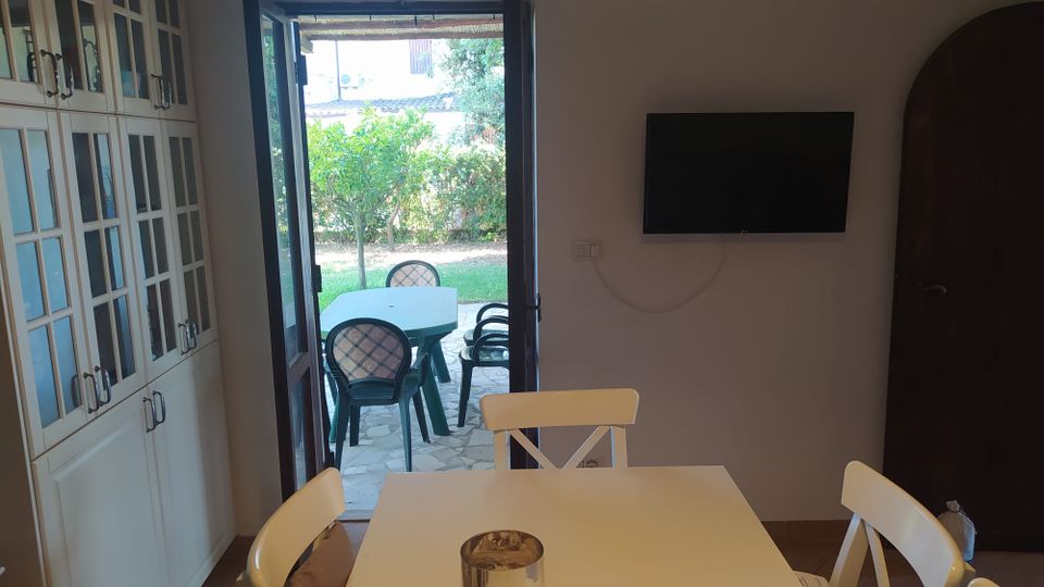 Sala da pranzo con tavolo bianco e sedie, armadi a vetri lungo la parete sinistra e TV a schermo piatto montata a destra. Una porta finestra aperta conduce a un patio con tavolo e sedie in plastica e affaccio su un giardino con prato e alberi.