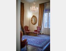 Salotto elegante con due poltrone in pelle rossa, grande tappeto decorato e pavimento in parquet. Lampadario in cristallo e specchio ovale dorato sulla parete, con alte finestre a portafinestra incorniciate da tende pesanti.