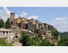 Borgo storico su un crinale con case in pietra addossate tra loro e una torre medievale prominente. Il complesso domina una valle verde con alberi e colline sullo sfondo sotto un cielo parzialmente nuvoloso.