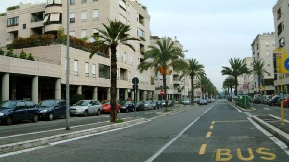 Ampio viale urbano con palme lungo lo spartitraffico e file di auto parcheggiate ai lati, fiancheggiato da alti edifici residenziali moderni. In primo piano si vede una corsia riservata ai bus con la scritta “BUS” sull’asfalto e segnaletica stradale.