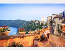 Terrazza panoramica su una strada acciottolata con muretto in pietra e vasi di fiori, affacciata su un lago circondato da colline boscose. Sul lato destro si vedono edifici del borgo arroccato e un lampione acceso lungo la via.