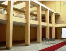 Cortile interno di un edificio con grandi colonne e ballatoi su più livelli, con pareti color beige e ringhiere metalliche. Nello spazio a terra ci sono alcune sedie, un tappeto rosso e uno schermo di proiezione su un lato.