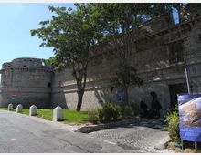 Mura di una fortezza in pietra con bastione cilindrico, affiancate da una strada asfaltata e da un’area verde con alberi e dissuasori in cemento. Accanto a un ingresso ad arco si vedono un’aiuola, una scultura scura e un cartello informativo.