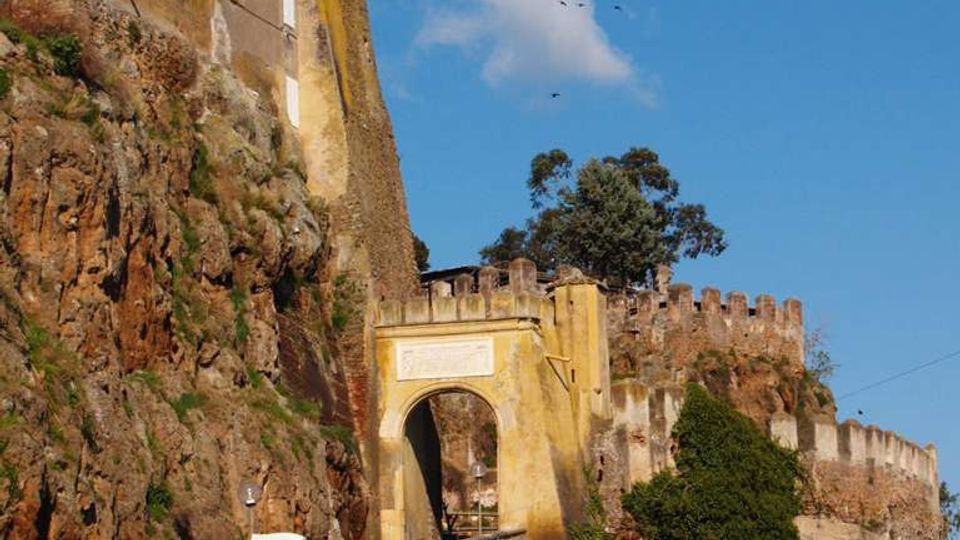 Fortezza in pietra costruita su una scogliera rocciosa, con edificio alto e mura merlate che seguono il profilo del promontorio. In primo piano si vede un arco d’ingresso giallo collegato a una strada in salita, con vegetazione sulle rocce e cielo azzurro sullo sfondo.