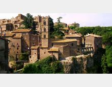 Borgo storico in pietra arroccato su uno sperone roccioso, con case addossate dai tetti in coppi e una torre campanaria centrale con finestre ad arco. Sullo sfondo si vede una collina boscosa e un ponte ad arcate che collega alcuni edifici sul margine del paese.