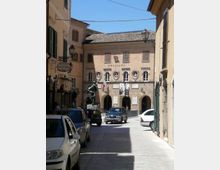Via del centro storico che conduce a una piazzetta con un palazzo in stile classico, facciata chiara con portici ad archi e finestre ovali decorative. Lampioni in ferro alle pareti, bandiera italiana e striscioni sulla facciata, con auto parcheggiate lungo la strada.