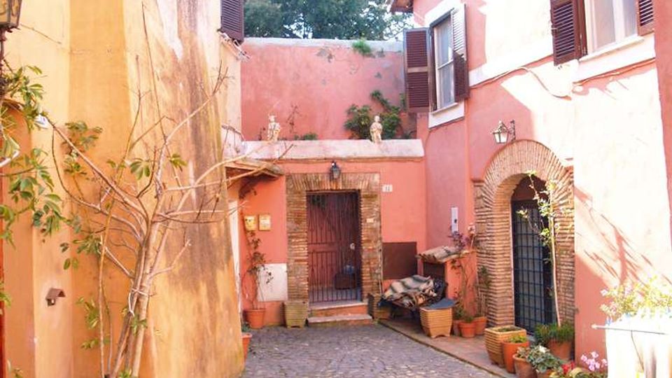 Cortile interno acciottolato tra edifici dai muri color ocra e rosa, con finestre a persiane e portoni ad arco in mattoni. Lungo le pareti ci sono vasi e fioriere con piante, e sul fondo un ingresso con cancello sotto una piccola tettoia.