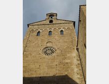 Facciata in pietra di una chiesa storica vista dal basso, con rosone circolare scolpito, tre finestre ad arco e una fila di archetti decorativi sotto il timpano. In cima si trova un piccolo campanile a vela con campana, sullo sfondo di cielo nuvoloso.