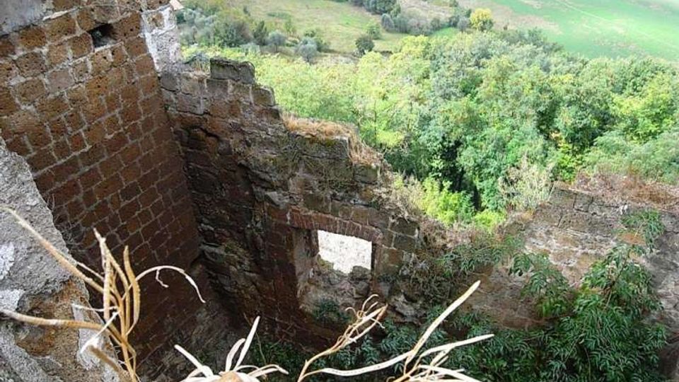 Rovine di una struttura in muratura di mattoni e pietra, con pareti parzialmente crollate e aperture rettangolari. Sullo sfondo si vede una vallata con boschi e campi coltivati.