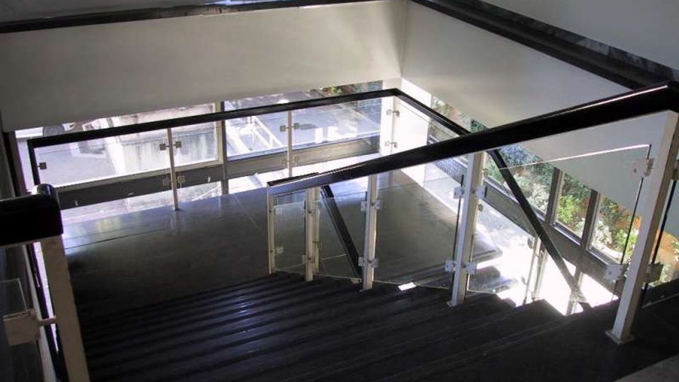 Interno di un vano scala moderno con gradini scuri e parapetti in vetro e metallo, affacciato su grandi finestre che lasciano entrare molta luce naturale. Le vetrate permettono la vista dell’esterno e di un’area con vegetazione.