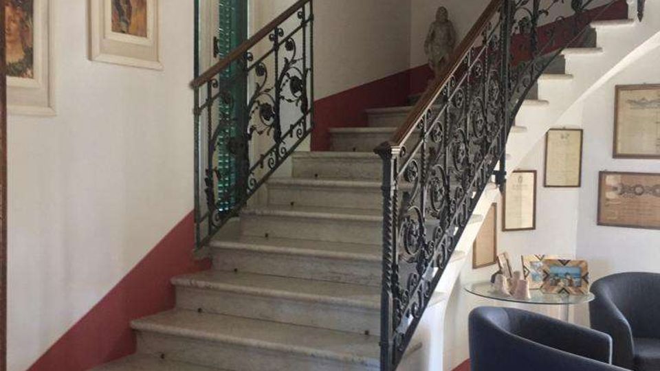 Interno di un edificio con ampia scalinata in marmo che curva verso l’alto, con ringhiere in ferro battuto decorato. Pareti bianche con fascia rossa, quadri e cornici alle pareti, una finestra con persiane verdi e un piccolo angolo salotto con poltrone e tavolino.