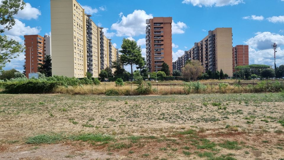 Ampia area erbosa e incolta in primo piano, delimitata da una recinzione, con sullo sfondo un complesso di alti palazzi residenziali a blocchi e balconi. Alberi sparsi e cielo azzurro con nuvole bianche completano la scena urbana.