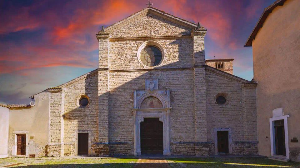 Facciata di una chiesa in pietra su una piccola piazza, con grande finestra circolare centrale e portale monumentale incorniciato. Ai lati si vedono edifici adiacenti e un cielo al tramonto con nuvole rossastre.