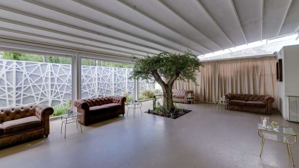 Ampio lounge interno con soffitto a travi bianche e grandi vetrate su tre lati, affacciate su un patio con pannelli geometrici. Al centro c’è un albero in una fioriera, con divani Chesterfield in pelle e piccoli tavolini metallici disposti lungo le pareti e davanti a tende chiare.