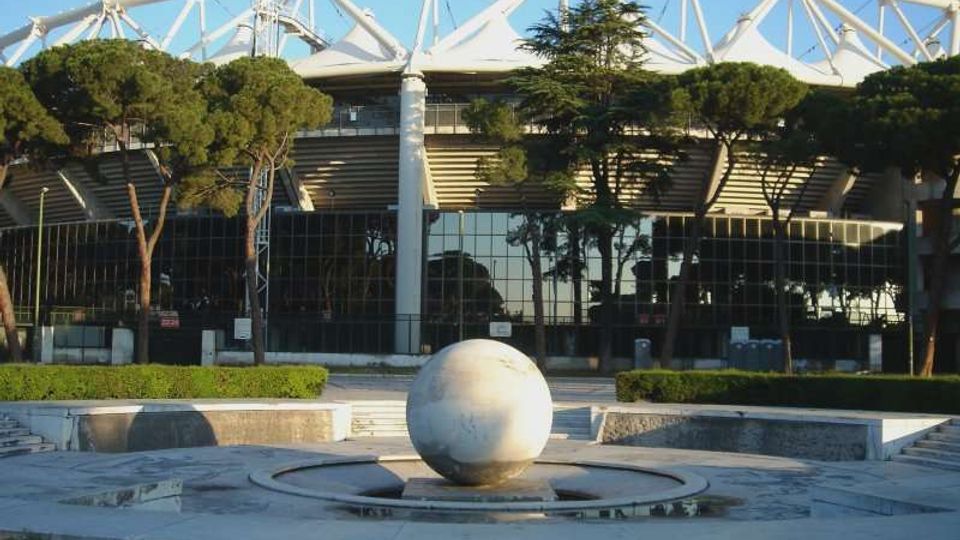 Veduta esterna di un grande stadio moderno con struttura bianca a travi e copertura a membrana, parzialmente nascosto da alberi. In primo piano si vede una piazza con una fontana circolare e una grande sfera in pietra al centro.