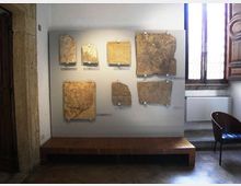 Pannello espositivo in una sala museale con grandi frammenti di pietra o lastre antiche fissati alla parete sopra una pedana in legno. Accanto si vede una finestra alta con inferriate e una parete in pietra, con pavimento in piastrelle.