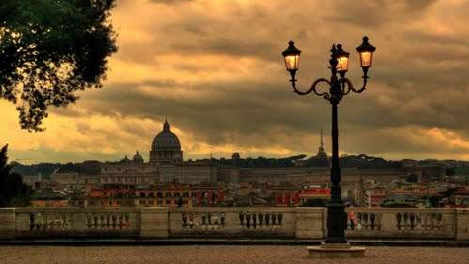 Terrazza panoramica con balaustra in pietra che affaccia sui tetti di Roma, con in primo piano un lampione in ferro a tre lanterne e un albero sul lato sinistro. Sullo sfondo si distingue la cupola della Basilica di San Pietro sotto un cielo nuvoloso al tramonto.