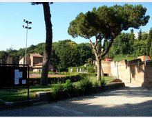 Area verde tipo parco con vialetto in ghiaia, siepi curate e grandi pini mediterranei. Sullo sfondo si vedono edifici bassi e una parete in mattoni lungo il percorso, con un palo dell’illuminazione e un pannello informativo.