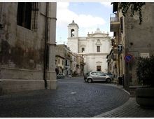 Strada acciottolata in un centro storico che conduce a una chiesa con facciata barocca chiara e campanile sullo sfondo. Ai lati si vedono edifici in pietra e intonaco, balconi e segnaletica stradale.