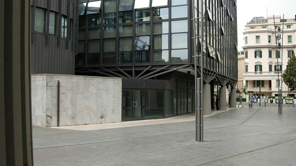 Ingresso di un moderno edificio per uffici con ampia facciata in vetro e struttura metallica, affacciato su una piazza pavimentata in cemento con pali di illuminazione. Sullo sfondo si vedono palazzi residenziali più tradizionali lungo la strada.