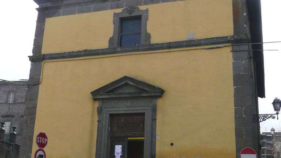 Facciata di una piccola chiesa o cappella con intonaco giallo e cornici in pietra scura, portale con timpano e finestra superiore sotto un frontone triangolare con nicchia. L’edificio si affaccia su una strada urbana con segnaletica stradale e un furgone parcheggiato davanti.