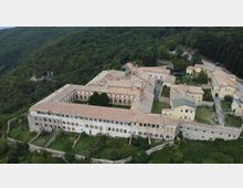Veduta aerea di un grande complesso monastico in pietra con tetti in coppi rossi e chiostro interno, disposto attorno a cortili e passaggi ad arcate. L’edificio è circondato da un muro di cinta e immerso in un paesaggio collinare boscoso.