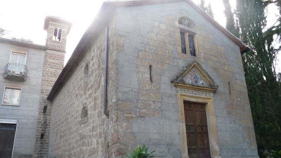 Piccola chiesa in pietra con facciata a capanna, portale in legno incorniciato da un arco e timpano decorato, e una stretta apertura verticale sopra l’ingresso. L’edificio è affiancato da un campanile in muratura e circondato da alberi alti, con un’area erbosa e vasi di piante sul davanti.