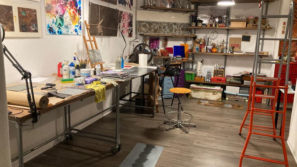 Interno di uno studio d’arte con grandi tavoli da lavoro su ruote, illuminazione a sospensione e pareti coperte da stampe e dipinti. Scaffalature sul fondo con materiali e strumenti artistici, sgabello centrale e scala rossa a destra su pavimento in legno scuro.