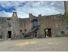 Cortile interno di una fortezza in pietra con alte mura merlate, un piccolo campanile e un arco sopraelevato raggiunto da una scalinata esterna. Sul lato destro si vede un grande portale aperto e, in alto, un camminamento con ringhiera sotto un cielo nuvoloso.