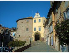 Veduta di una piazza in un borgo storico con edificio giallo a più piani e grande arco d’ingresso in pietra, affiancato da una torre cilindrica in muratura. Ai lati si vedono case con persiane e balconi fioriti, pavimentazione in pietra e lampioni, sotto un cielo sereno.