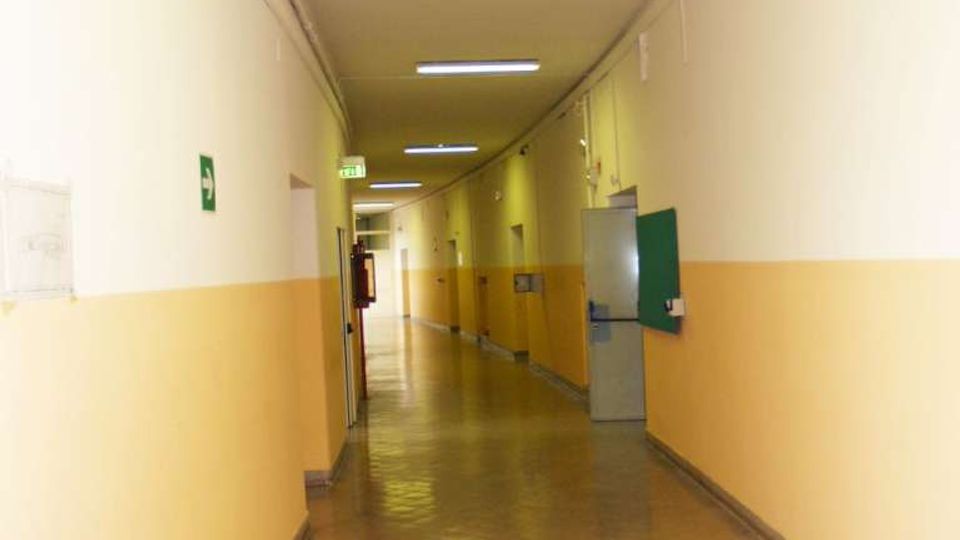 Lungo corridoio interno di un edificio, con pareti bianche e fascia inferiore gialla, pavimento lucido e illuminazione a neon sul soffitto. Ai lati si vedono porte e segnaletica di sicurezza, con un’uscita sul fondo del passaggio.