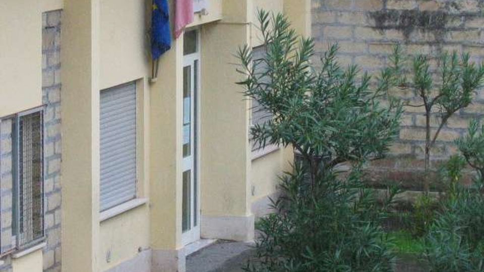 Facciata di un edificio pubblico color giallo chiaro con ingresso, finestre con tapparelle e una bandiera dell’Unione Europea e italiana vicino alla porta. In primo piano si vede un piccolo giardino con arbusti e un muretto di pietra sul lato destro.