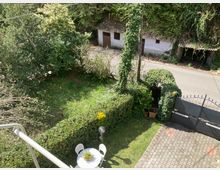 Vista dall’alto di un giardino privato con prato, siepi potate e tavolino con sedie vicino a un vialetto pavimentato e a un cancello metallico. Sullo sfondo si vede una strada e piccoli edifici rurali con tetto in tegole, circondati da alberi e vegetazione fitta.
