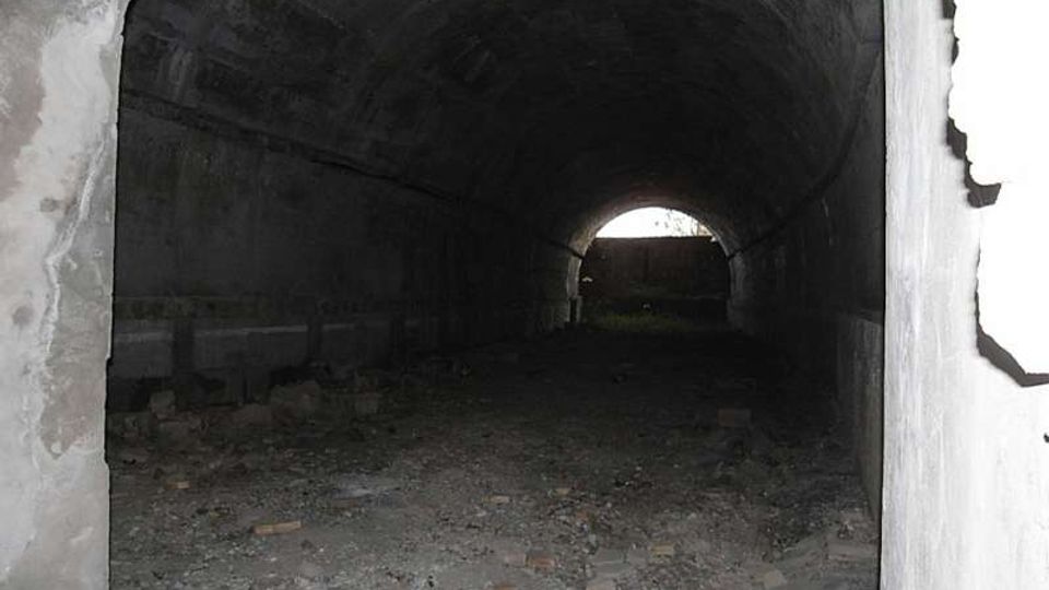 Interno di un tunnel o passaggio sotterraneo in cemento con volta a botte, pareti scure e pavimento coperto di macerie e detriti. In fondo si vede un’apertura ad arco che lascia entrare luce dall’esterno.