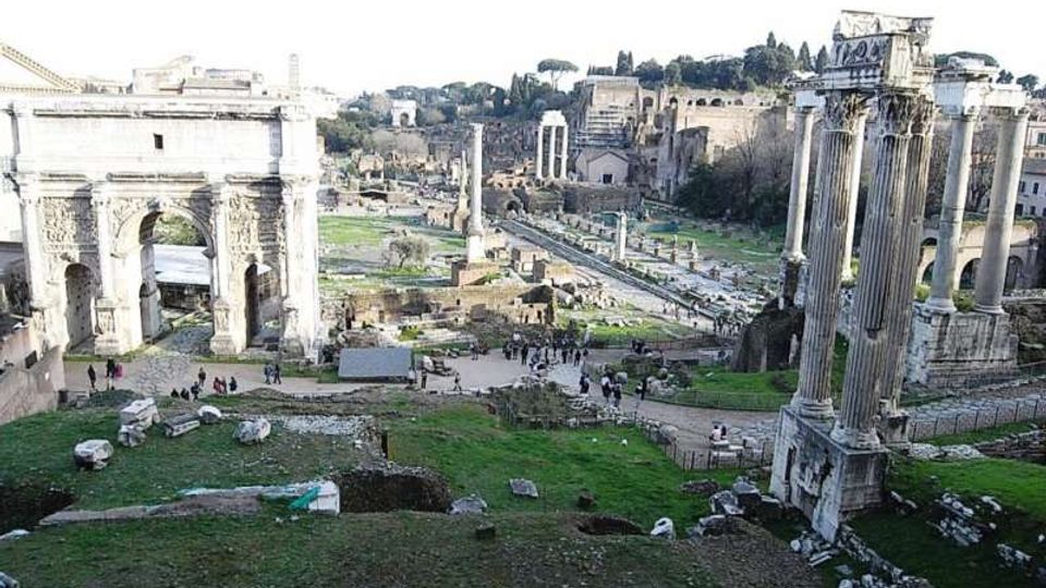 Veduta panoramica del Foro Romano con rovine antiche, includendo un grande arco in pietra sulla sinistra e alte colonne di un tempio sulla destra. Al centro si estende l’area archeologica con resti di edifici e percorsi tra prati e pietre, sullo sfondo delle colline e della città.
