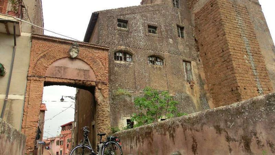 Antico complesso in mattoni con grande arco d’ingresso e torre merlata, affiancato da edifici in muratura con piccole finestre. In primo piano si vede un vicolo lastricato con un basso muro in pietra e due biciclette appoggiate.