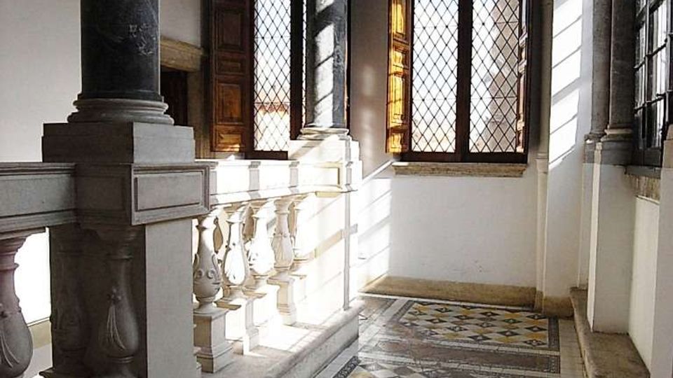 Loggia o corridoio interno di un edificio storico con colonne in pietra e balaustra scolpita, soffitto a cassettoni e grandi finestre con vetri a losanghe e scuri in legno. Pavimento in piastrelle decorate a motivi geometrici, illuminato dalla luce naturale che entra lateralmente.