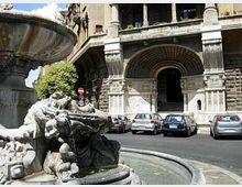 Fontana in pietra con sculture e vasca circolare in primo piano, in una piazza urbana. Sullo sfondo si vede un grande edificio storico in pietra con portico ad archi, colonne e un ingresso monumentale, con auto parcheggiate lungo la strada.