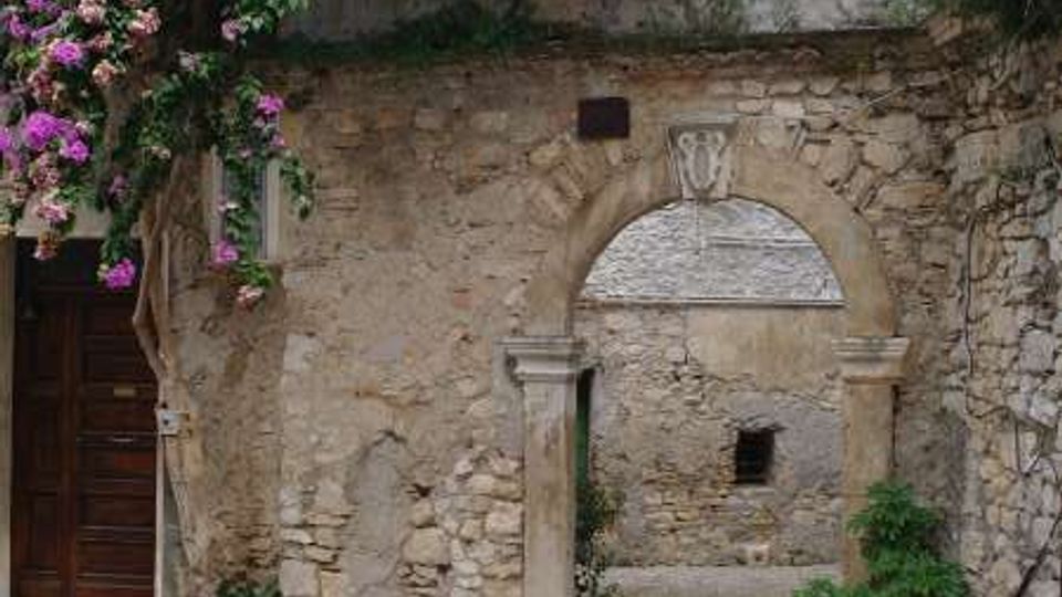Cortile in pietra con pavimentazione a ciottoli e un arco in muratura che conduce a un vicolo interno, affiancato da un vaso con pianta. Sopra l’arco, una fitta bougainvillea con fiori viola e chiari ricade dal balcone e copre parte della facciata.